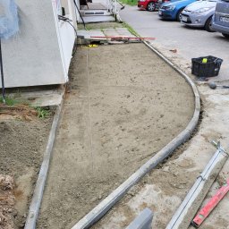 Smart Solutions - Przygotowanie podłoża pod chodnik z obrzeżami betonowymi, widoczna napięta linka wyznaczająca linię, poziomica, skrzynka z narzędziami i zaparkowane samochody w tle.