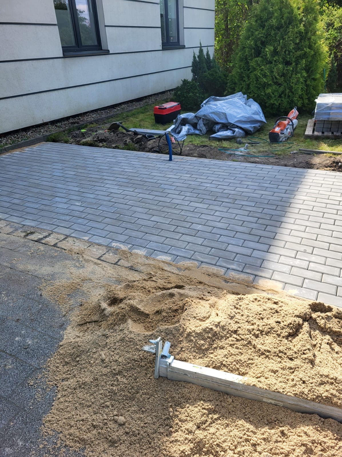 Fragment układanej szarej kostki brukowej, widoczny podsyp z piasku, narzędzia i maszyna do cięcia kostki w tle, dom z jasną elewacją i zielone tuje.