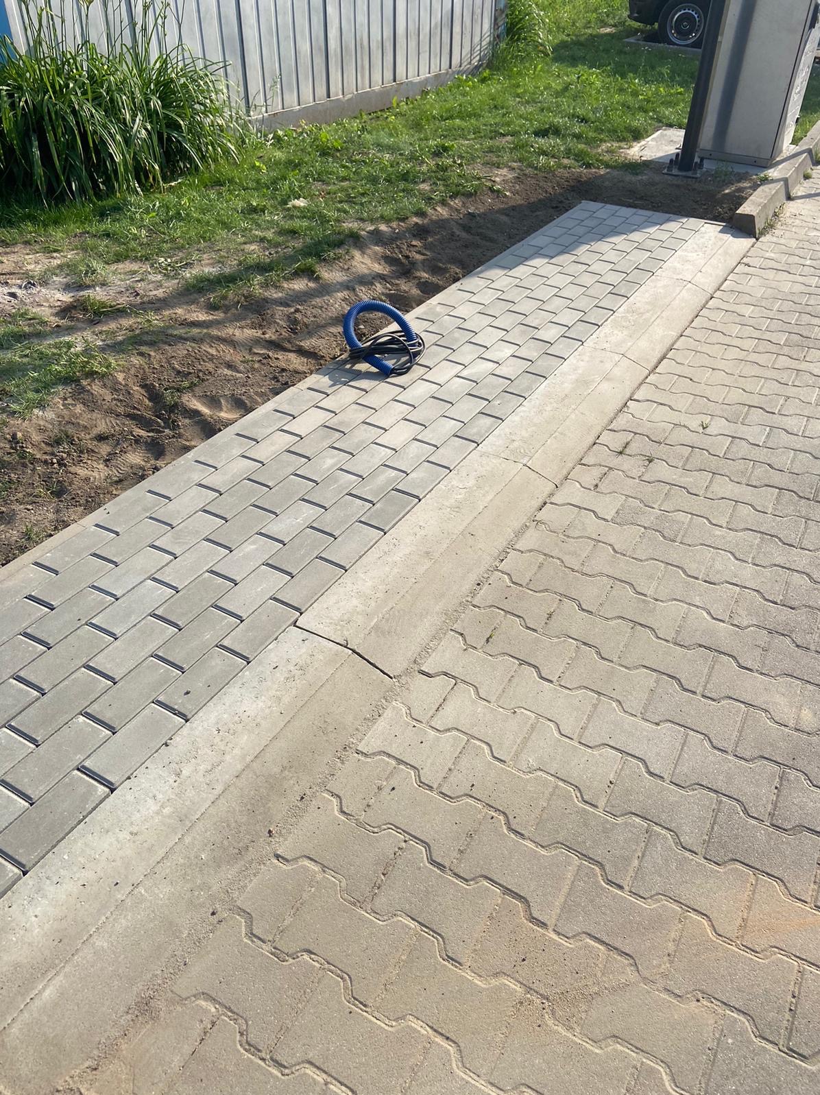 Świeżo ułożona, szara kostka brukowa sąsiaduje ze starą nawierzchnią, na skraju leży niebieski wąż karbowany.