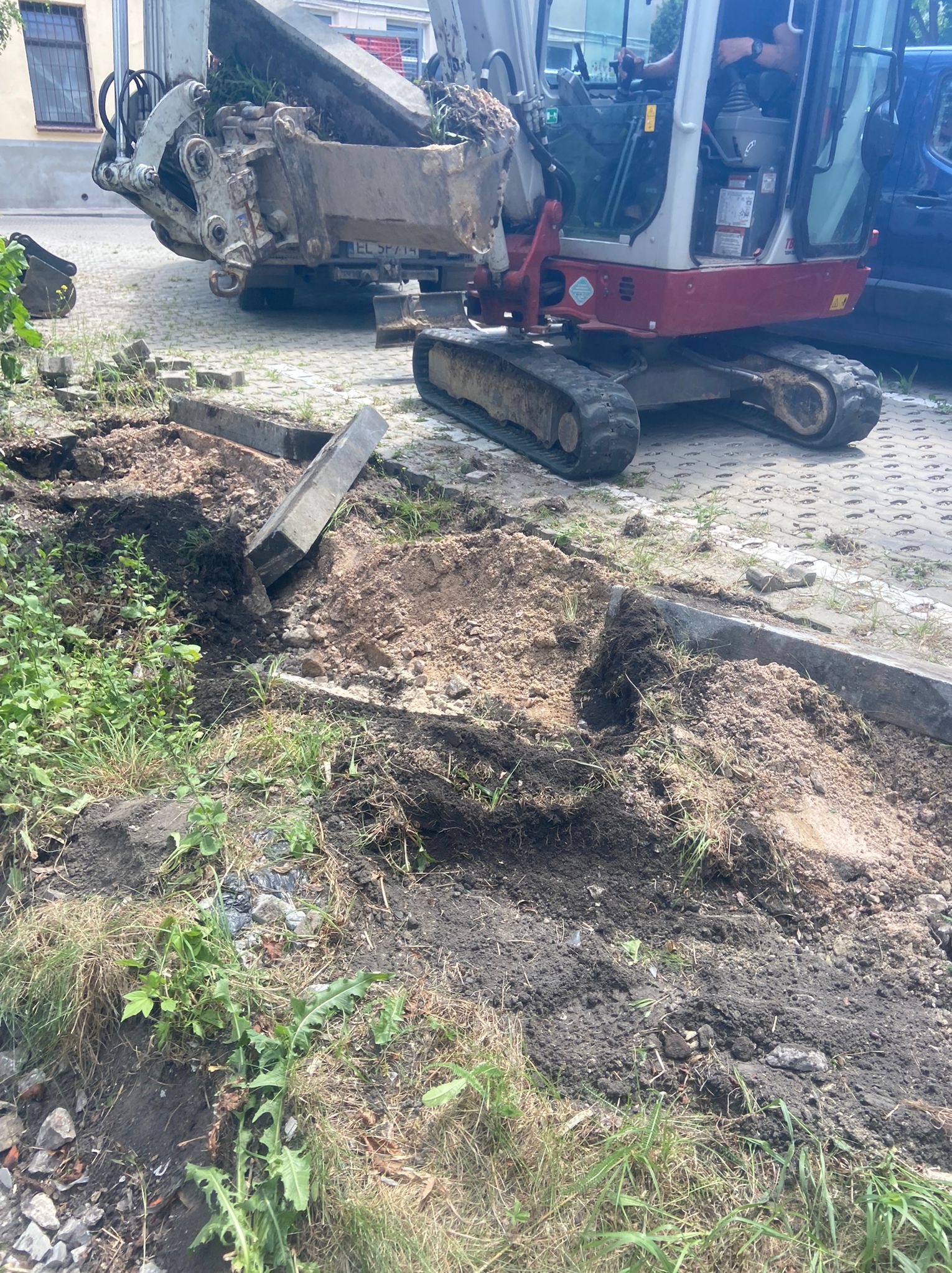 Mała koparka na gąsienicach podczas prac ziemnych przy krawężniku chodnika, z wykopanym wąskim rowem i stertą ziemi obok.