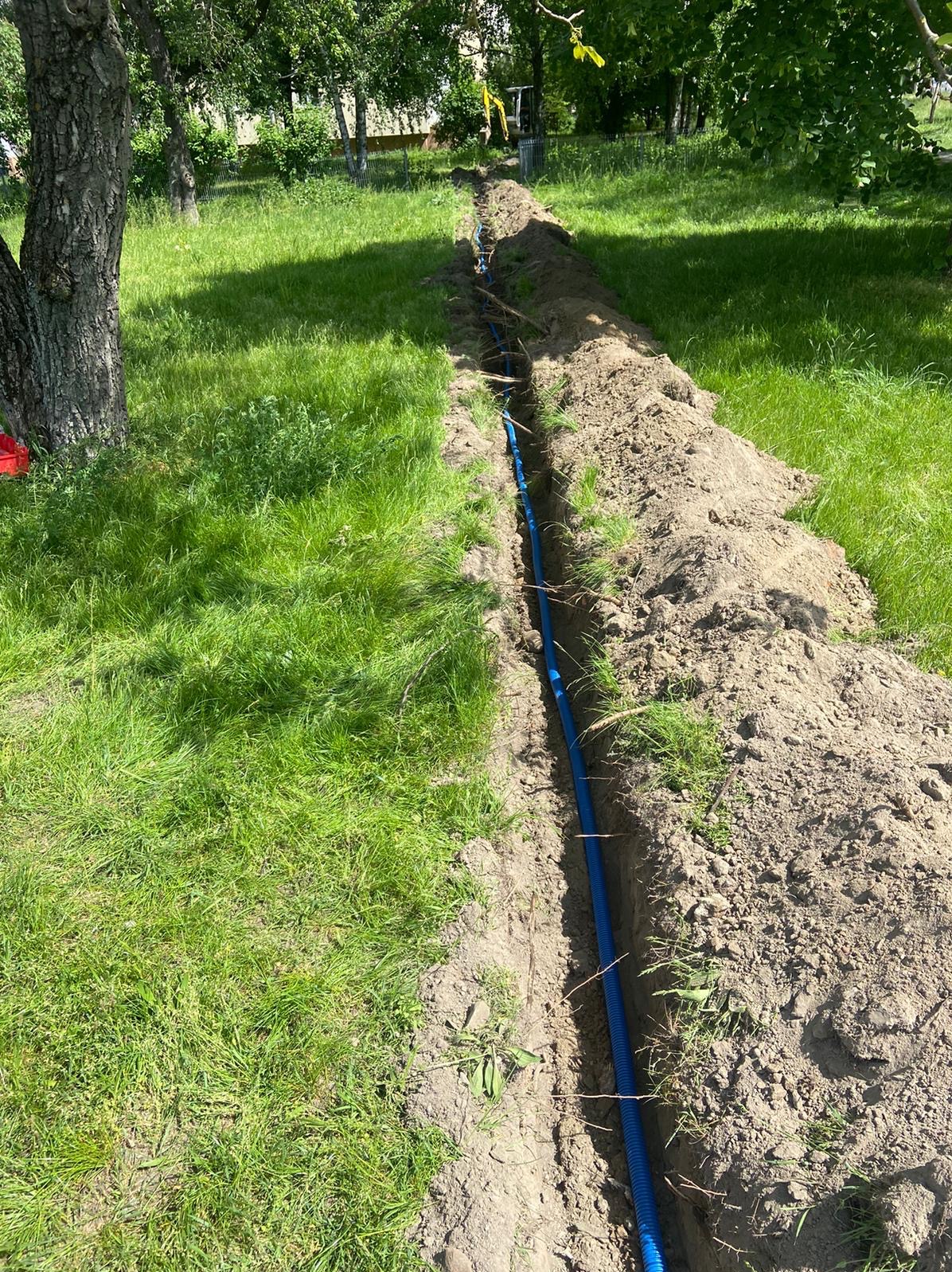 Wykopany wąski rów z niebieską rurą karbowaną ułożoną wzdłuż, na tle zielonej trawy i drzew.