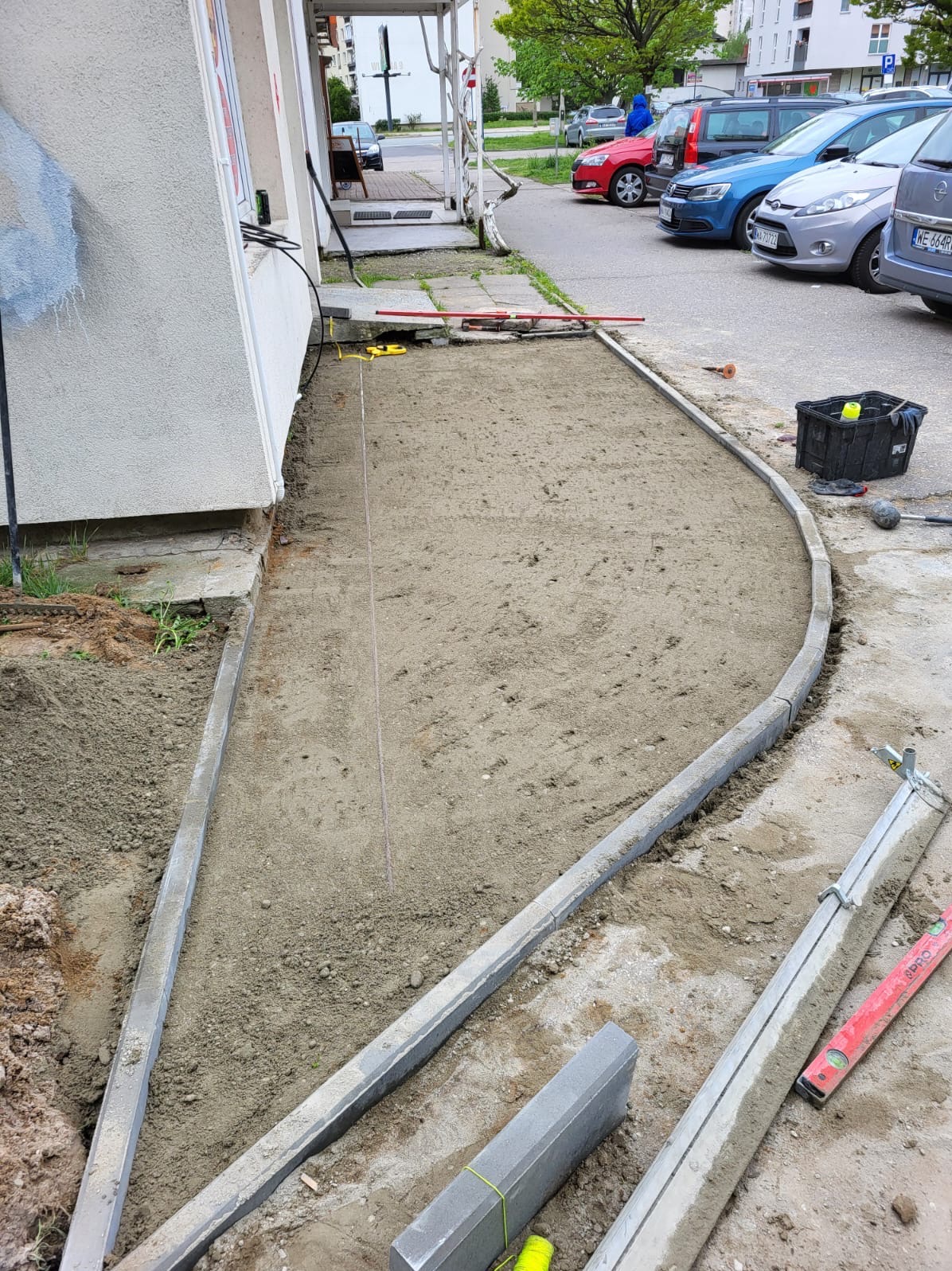 Przygotowanie podłoża pod chodnik z obrzeżami betonowymi, widoczna napięta linka wyznaczająca linię, poziomica, skrzynka z narzędziami i zaparkowane samochody w tle.