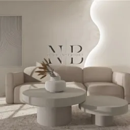 Beżowa sofa z dwoma okrągłymi stolikami kawowymi, wazonem z suchymi gałązkami i wazonem z zielonymi gałązkami na tle jasnego pokoju z abstrakcyjnym obrazem na ścianie i falistą dekoracją ścienną.