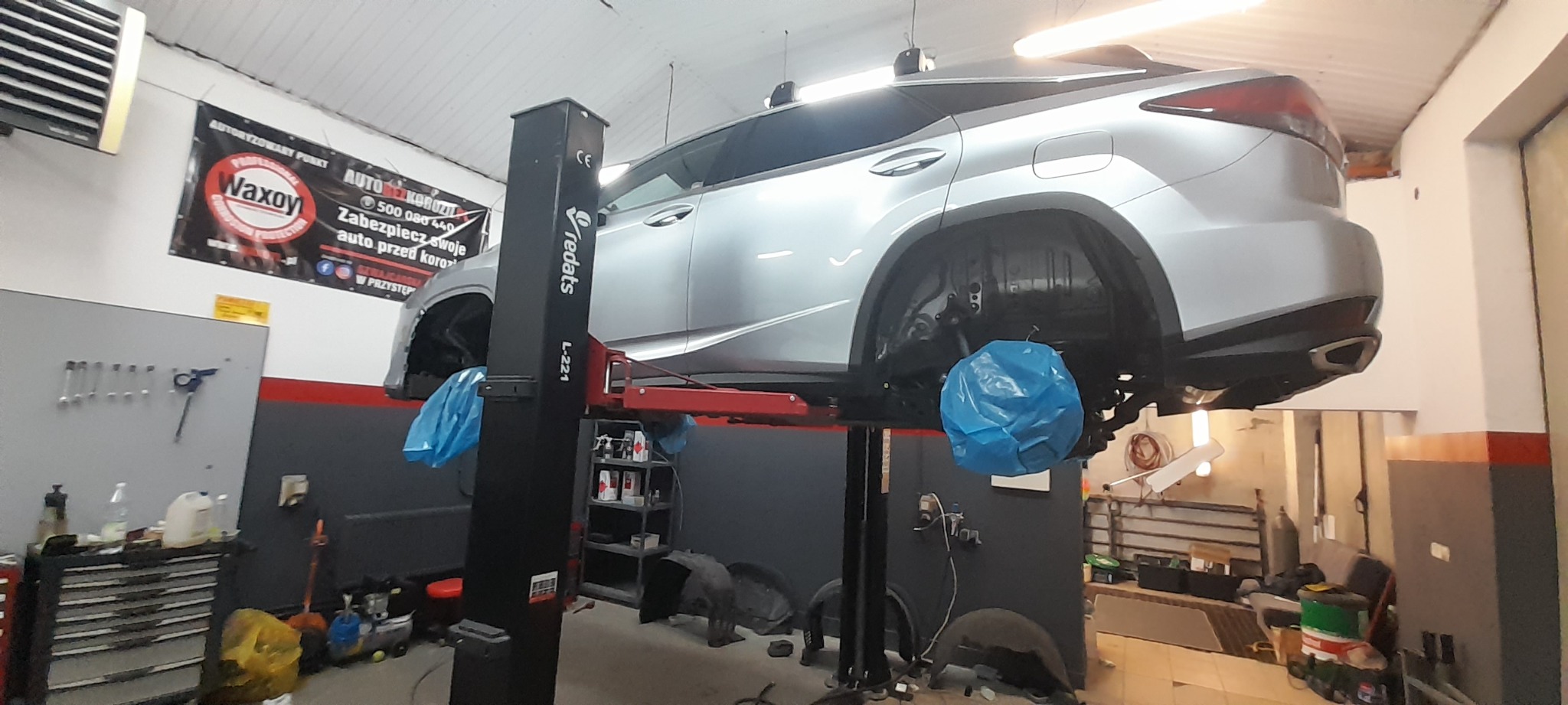 Srebrny SUV na podnośniku warsztatowym Redats L-221, z zabezpieczonymi elementami zawieszenia niebieską folią, w tle narzędzia i baner reklamowy Waxoy.