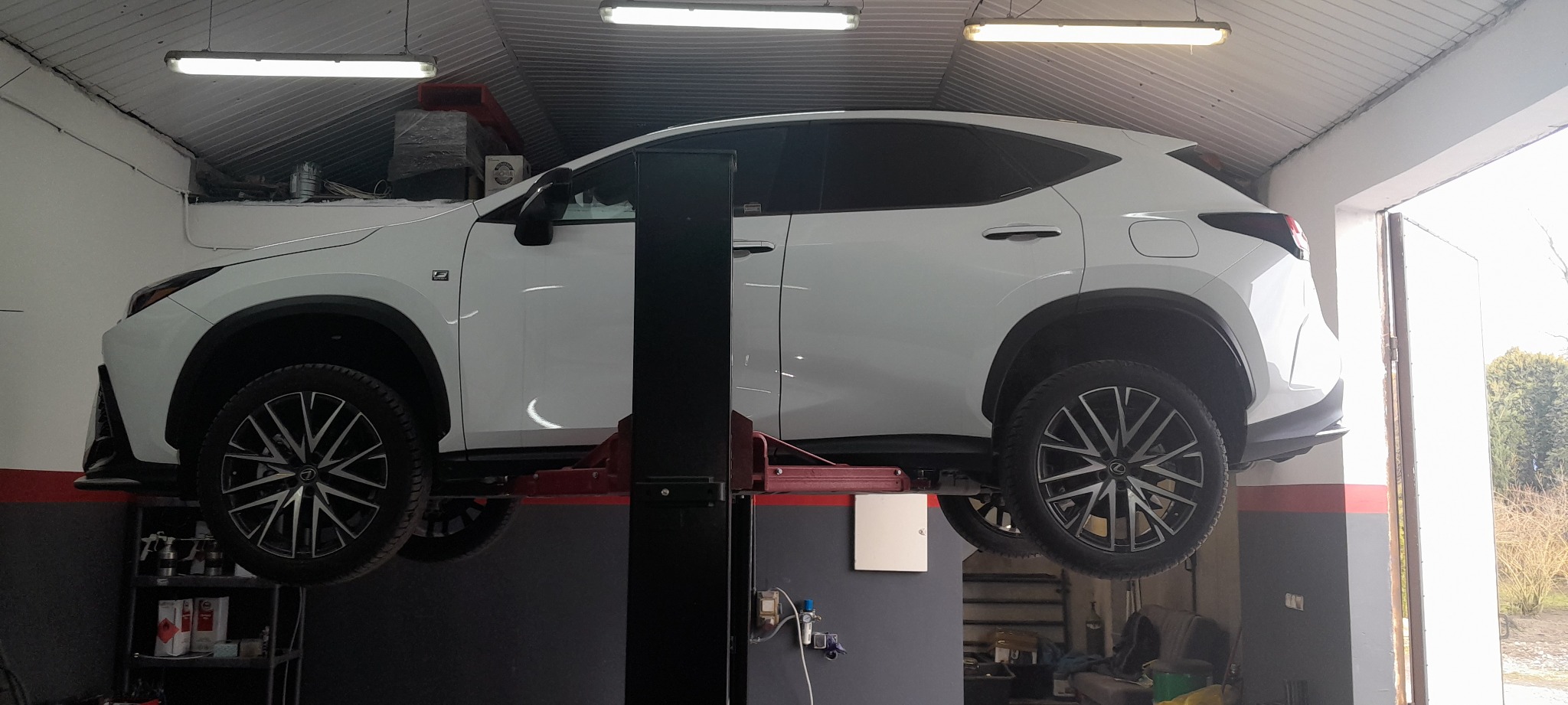 Biały samochód Lexus NX na podnośniku w warsztacie, widoczne podwozie i koła. Ściany warsztatu pomalowane na szaro z czerwonym pasem. W tle regał z narzędziami i preparatami.