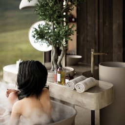 blank vis studio - Kobieta relaksuje się w wannie wypełnionej pianą, z widokiem na zielony krajobraz za oknem; obok wanny stoi umywalka z ręcznikami i kosmetykami, a całość dopełnia dekoracyjne drzewko w doniczce.