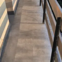 Wyłożony szarymi płytkami balkon z drewnianą balustradą z elementami pomalowanymi na czarno, na ścianie widoczne cienie od promieni słonecznych, na balustradzie zamontowana antena satelitarna.