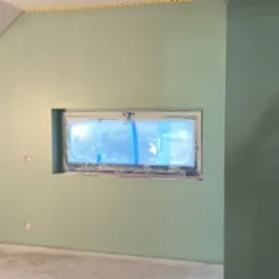 Ściany w odcieniach zieleni w pokoju na poddaszu, okno zabezpieczone folią malarską, widoczne ślady szpachlowania i taśmy malarskiej przy suficie.