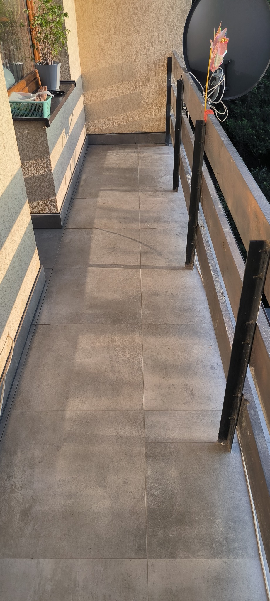 Wyłożony szarymi płytkami balkon z drewnianą balustradą z elementami pomalowanymi na czarno, na ścianie widoczne cienie od promieni słonecznych, na balustradzie zamontowana antena satelitarna.