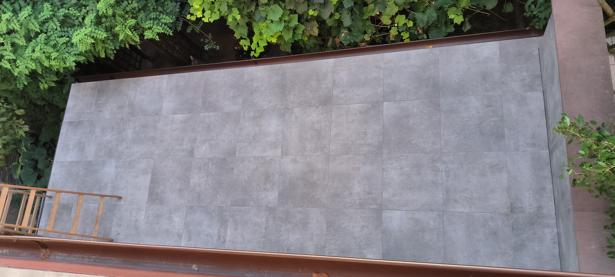 Wykończony taras z szarych, kwadratowych płyt, widok z góry, z zielenią roślin w tle i drewnianą drabiną opartą o ścianę.