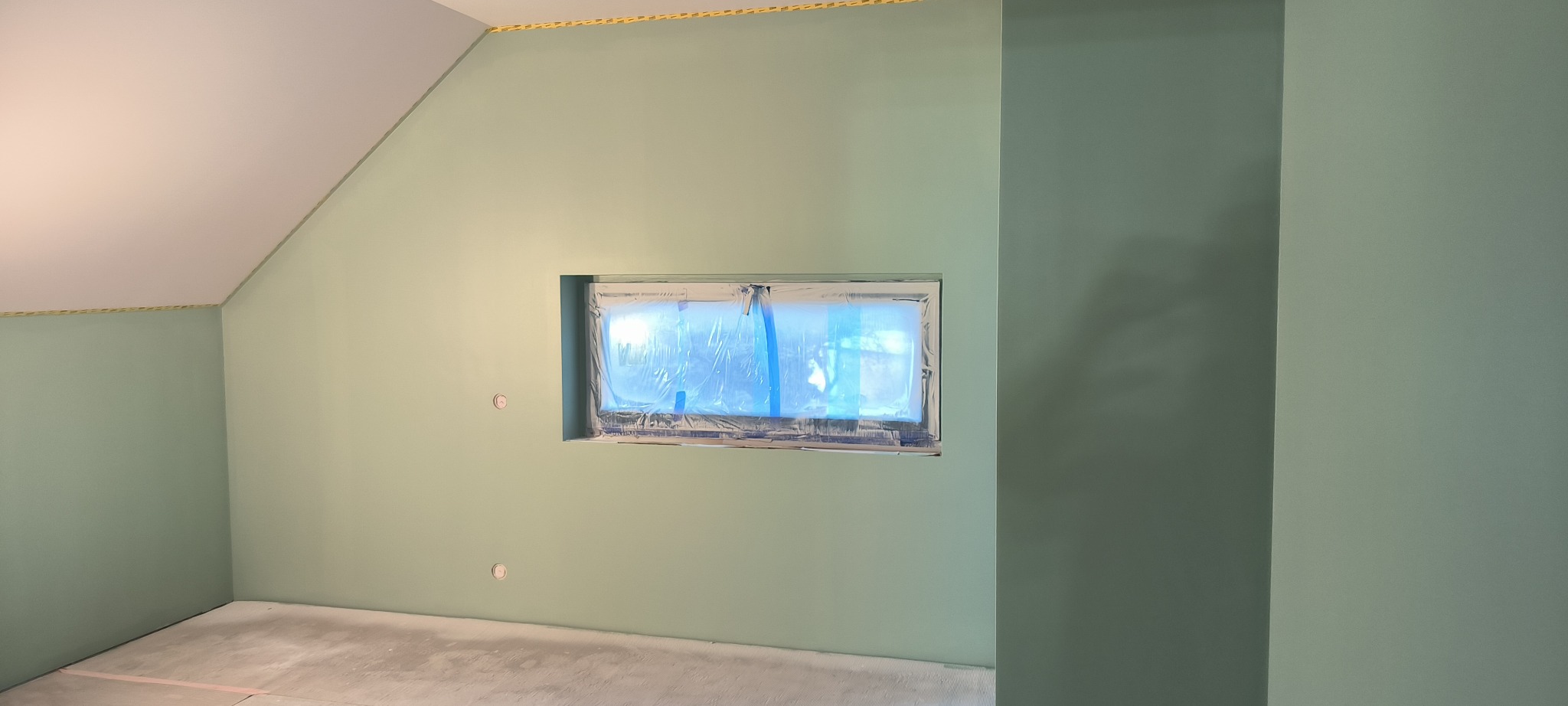 Ściany w odcieniach zieleni w pokoju na poddaszu, okno zabezpieczone folią malarską, widoczne ślady szpachlowania i taśmy malarskiej przy suficie.