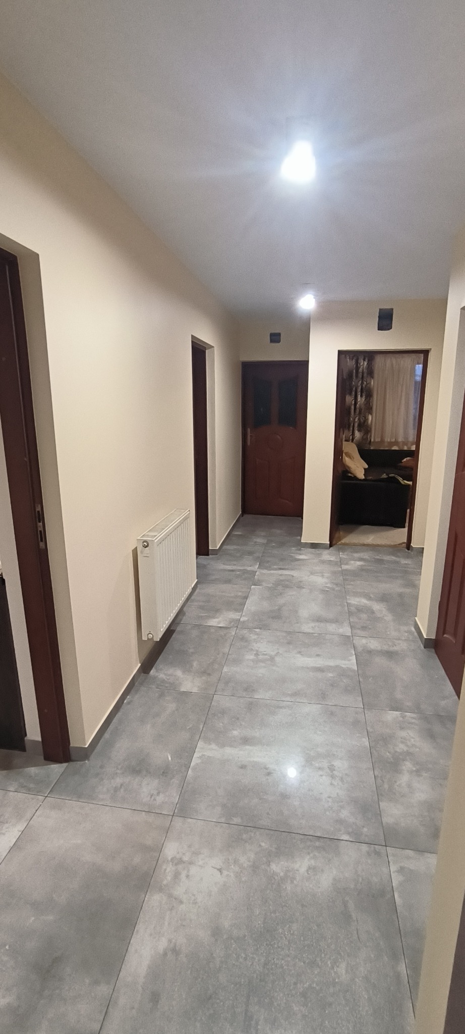 Korytarz po remoncie z szarymi płytkami podłogowymi imitującymi beton, kremowe ściany, biały grzejnik i ciemne drewniane drzwi po obu stronach.