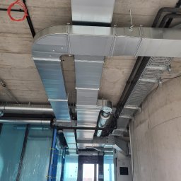 DMB Montage S.C. - Widok sufitu w budynku w Zielonej Górze w trakcie instalacji systemu wentylacyjnego z metalowymi kanałami i przewodami elektrycznymi, widoczna klatka schodowa w tle.