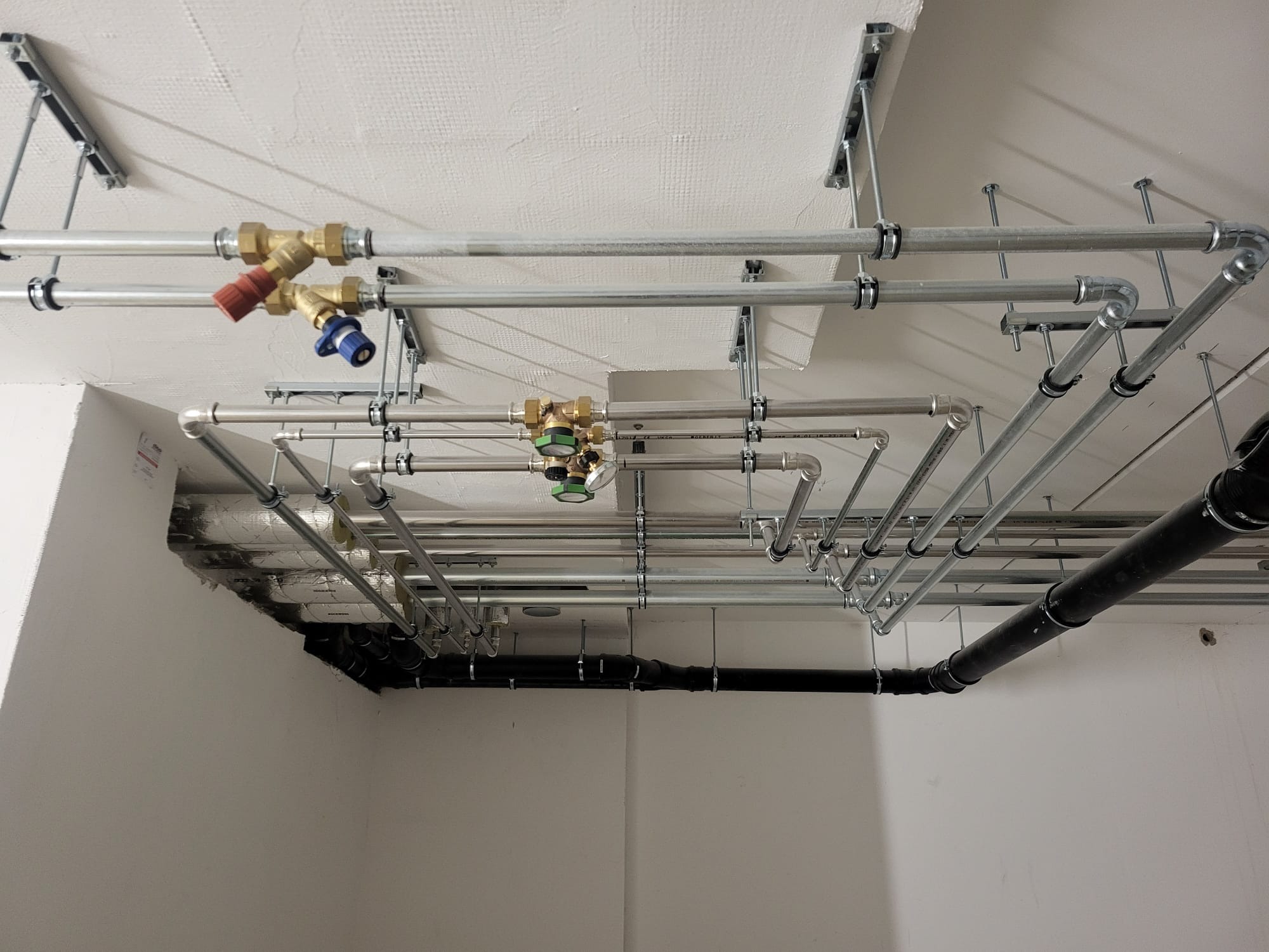 Skomplikowany system rur instalacyjnych z zaworami, zamocowany pod sufitem w jasnym pomieszczeniu. Widoczne rury stalowe, czarne rury kanalizacyjne oraz elementy mocujące.