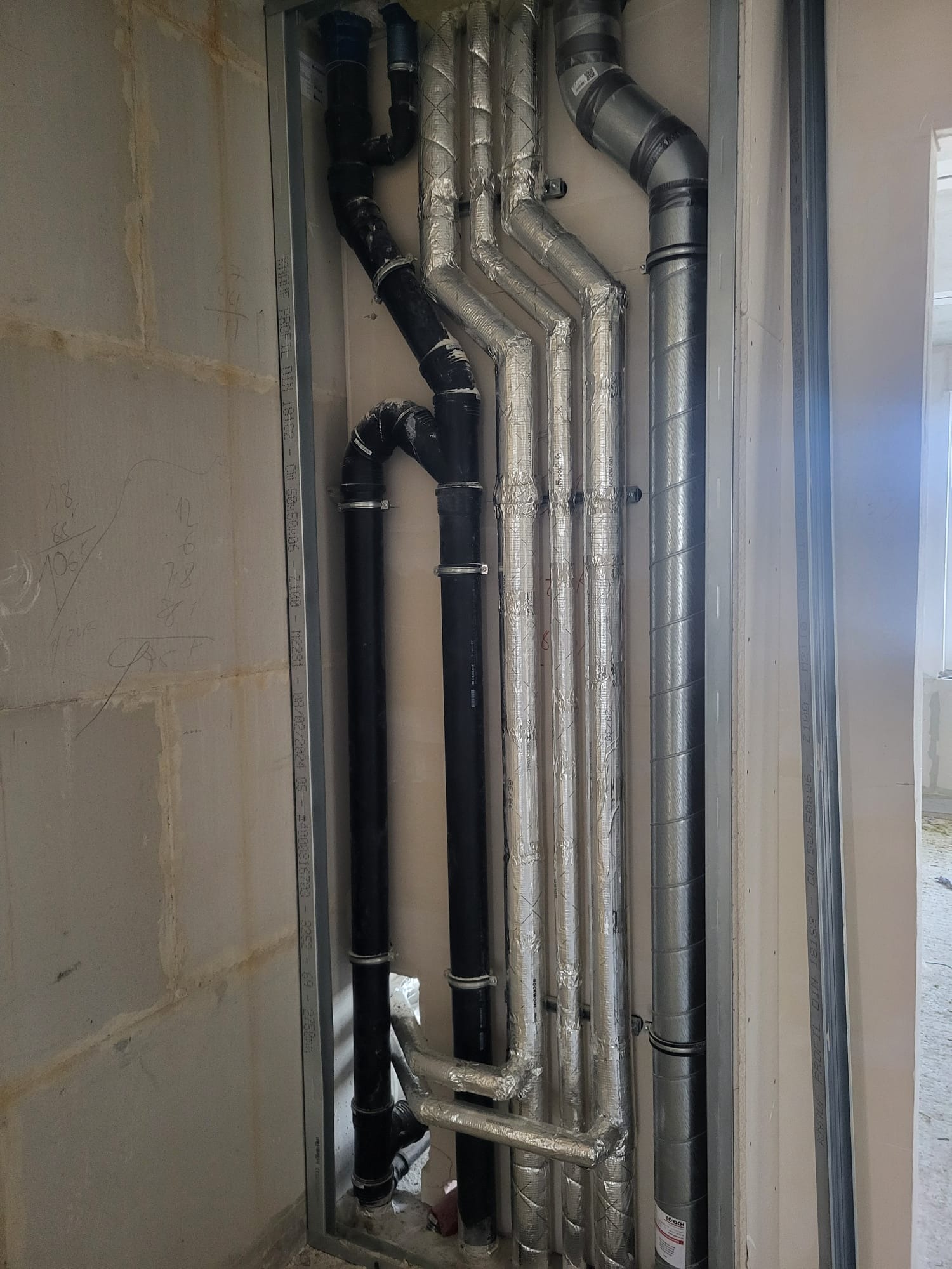 Surowe ściany z widocznymi rurami instalacji wodnej i grzewczej, niektóre owinięte folią izolacyjną, zamontowane w metalowej ramie w trakcie budowy.