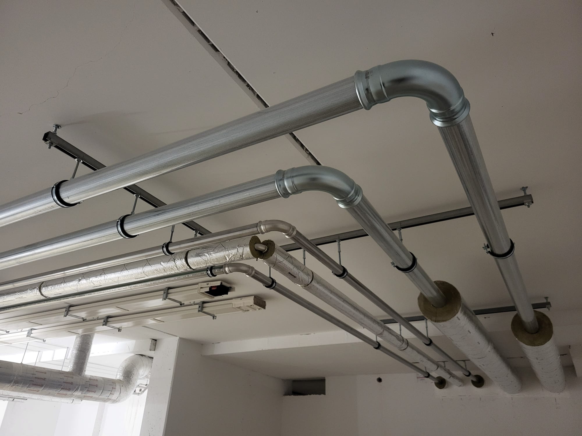 Metalowe rury pod sufitem, niektóre zaizolowane, zamocowane na metalowych wspornikach, widoczne złączenia i kolanka.