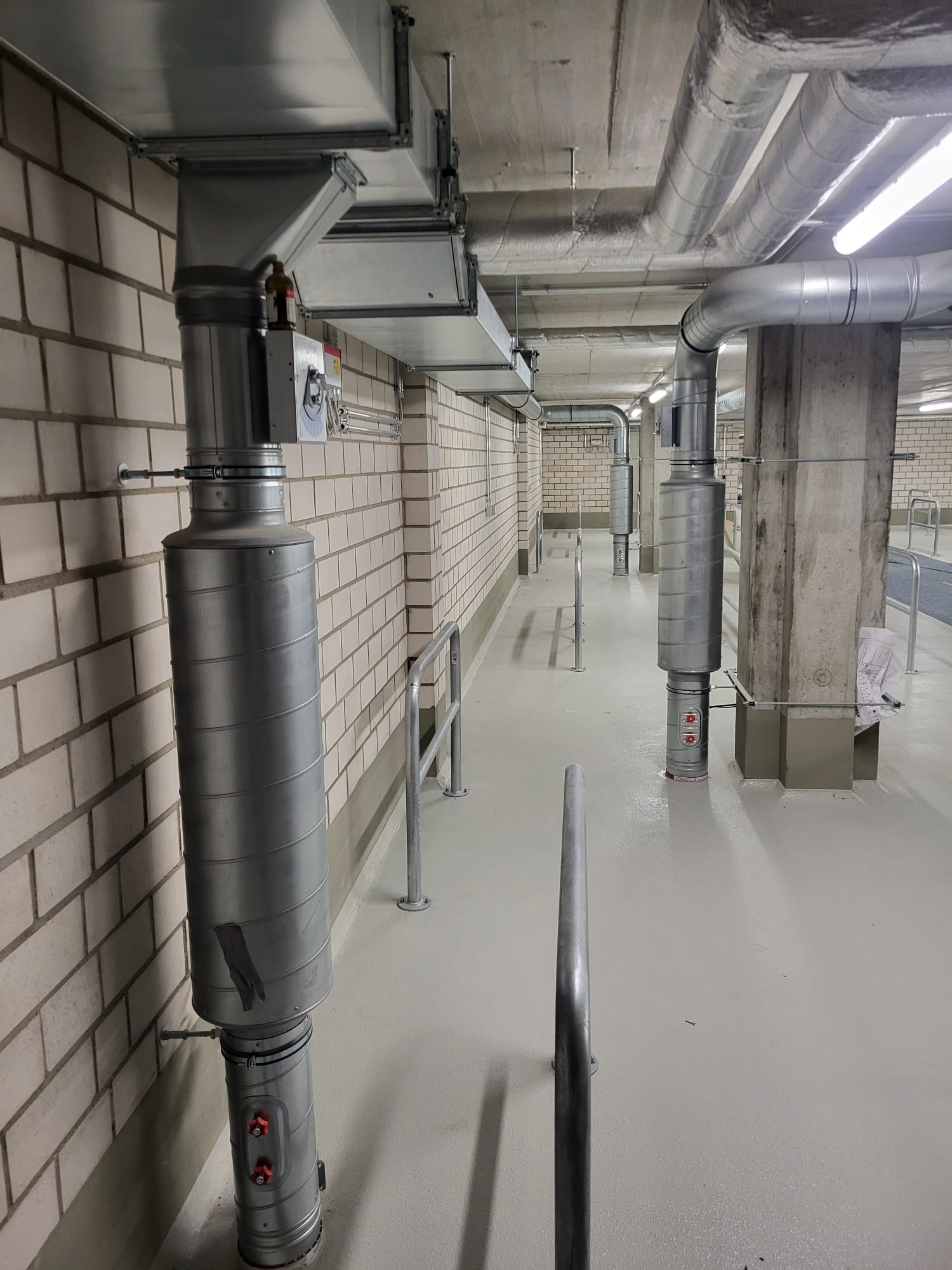 Szereg pionowych, metalowych rur wentylacyjnych z izolacją termiczną, przymocowanych do ceglanej ściany wzdłuż korytarza z betonowym sufitem i jasną posadzką.