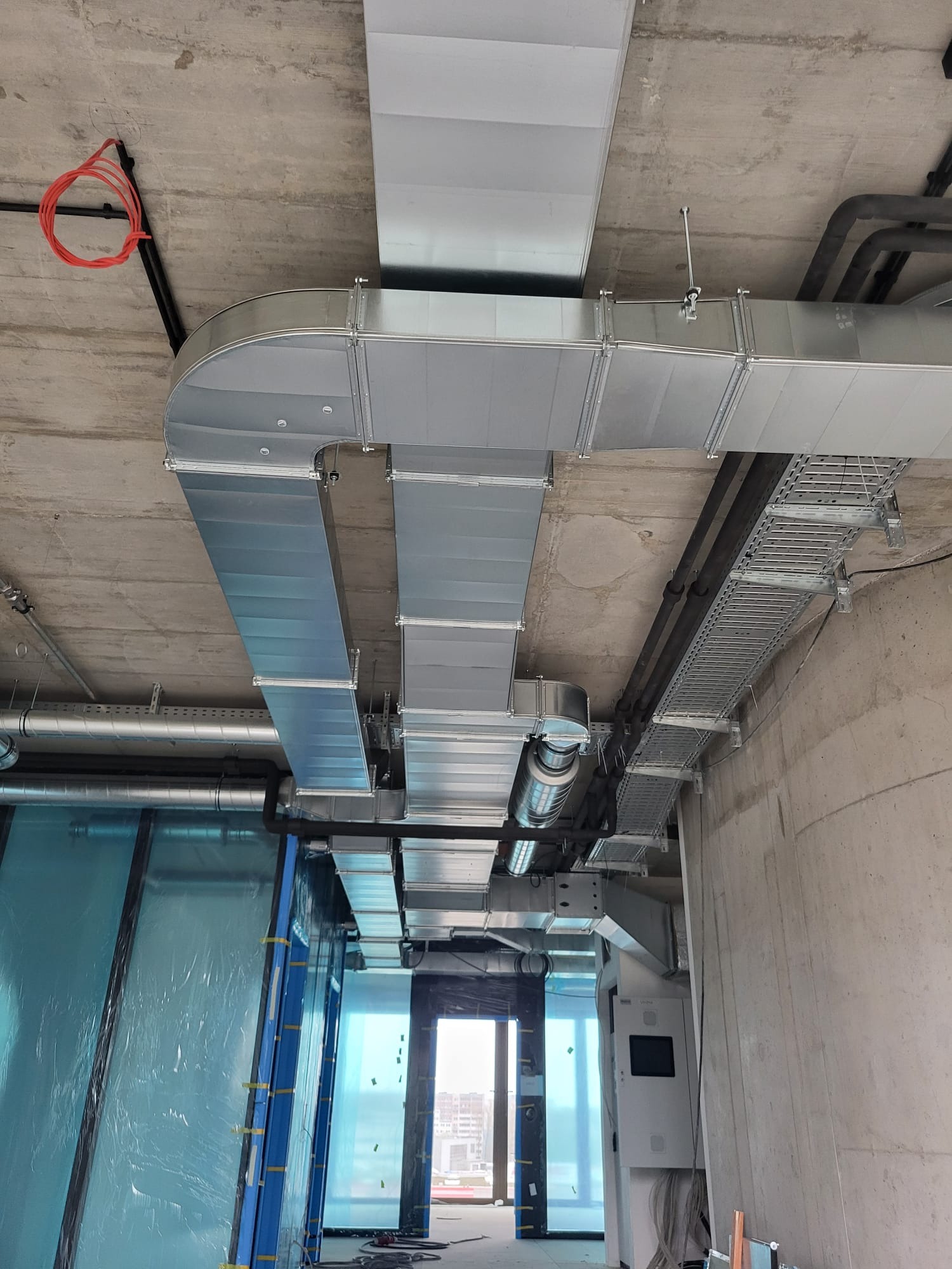 Widok sufitu w budynku w Zielonej Górze w trakcie instalacji systemu wentylacyjnego z metalowymi kanałami i przewodami elektrycznymi, widoczna klatka schodowa w tle.