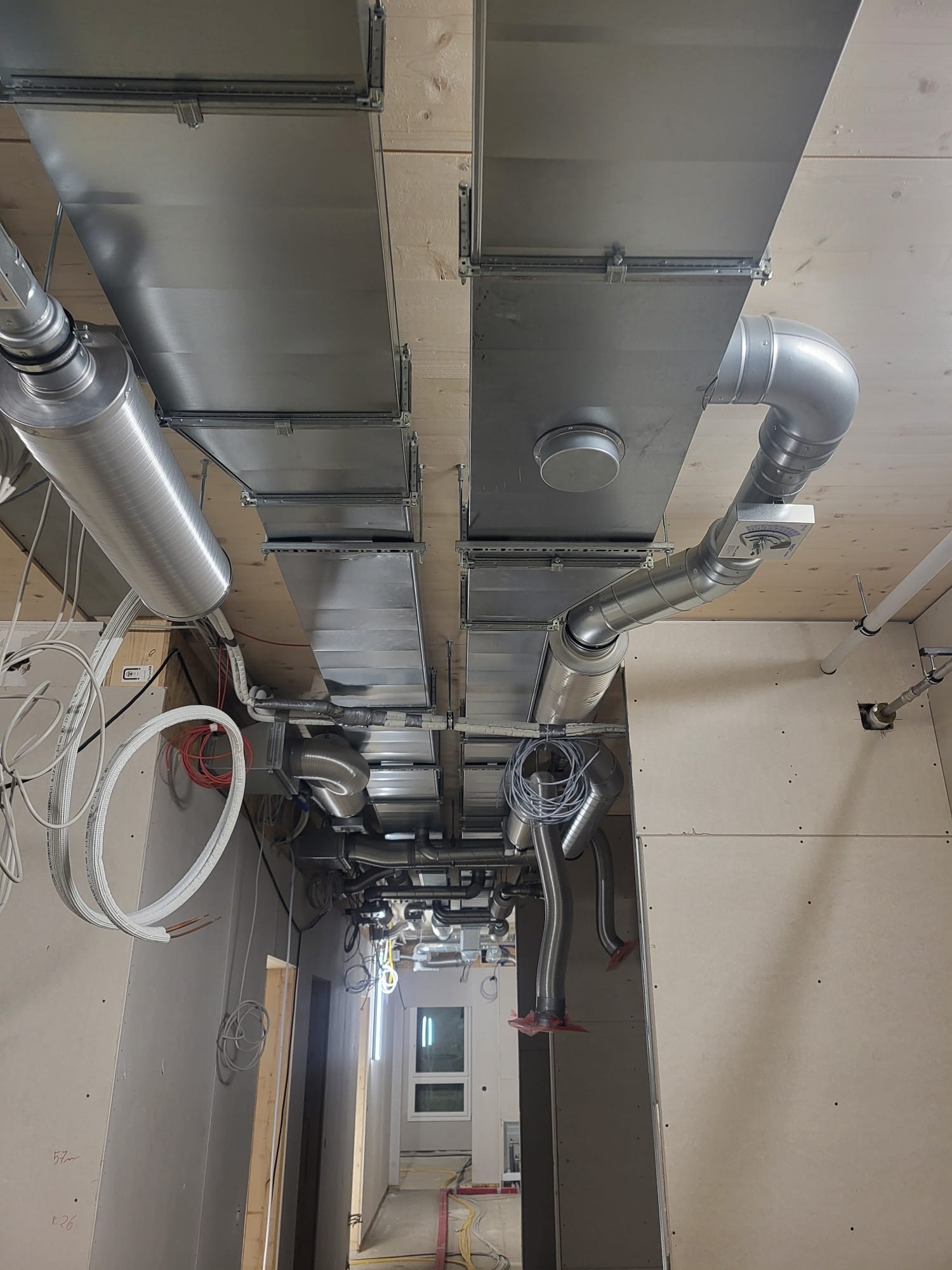 Widok na sufit z zamontowanymi systemami wentylacyjnymi z metalowych kanałów, rur i kształtek, w pomieszczeniu w stanie surowym, z widocznymi kablami i elementami konstrukcyjnymi.