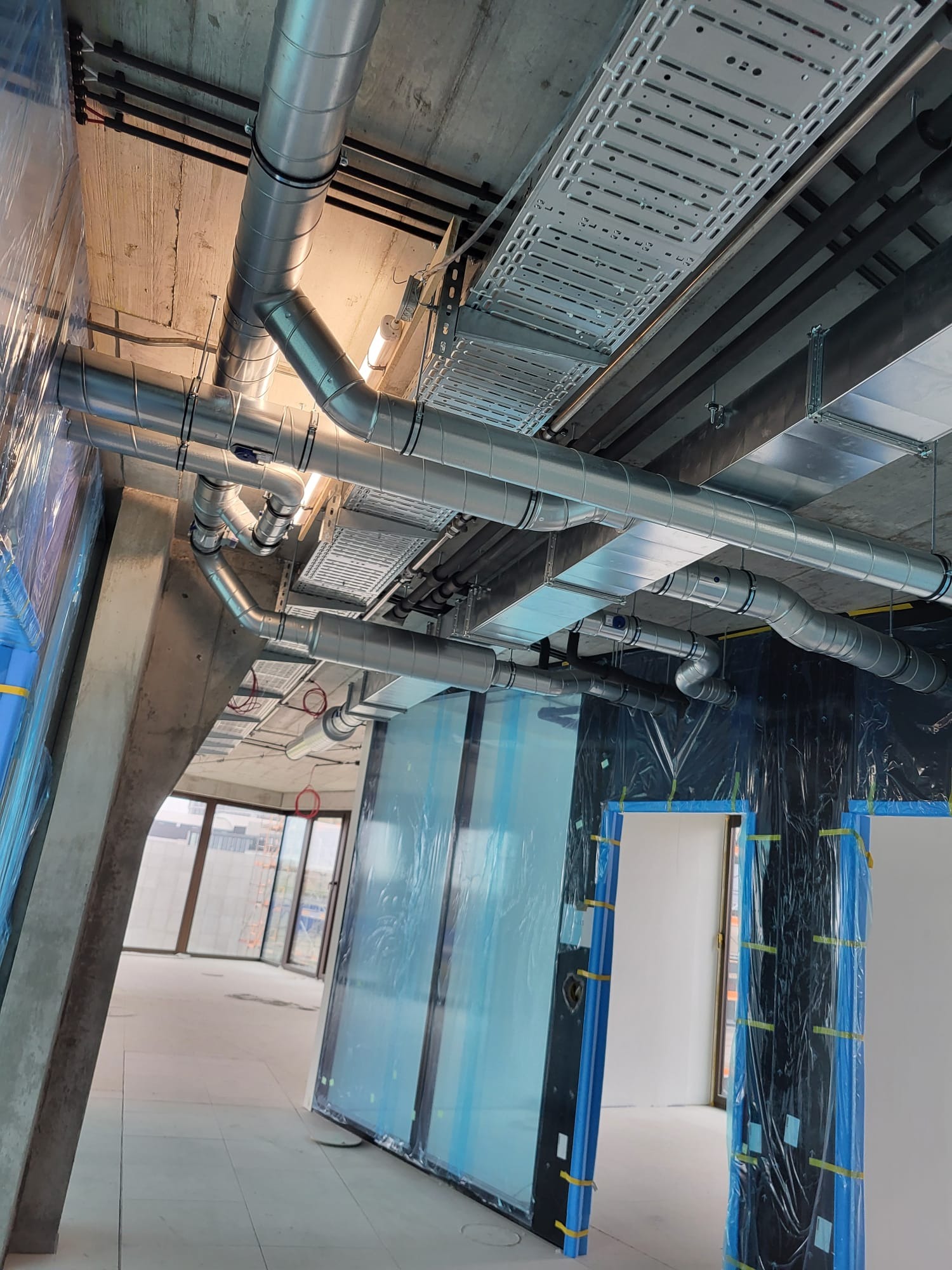 Widok z dołu na instalację wentylacyjną z rur spiro, koryta kablowe i betonowy strop w surowym stanie, zabezpieczone folią okna w tle.
