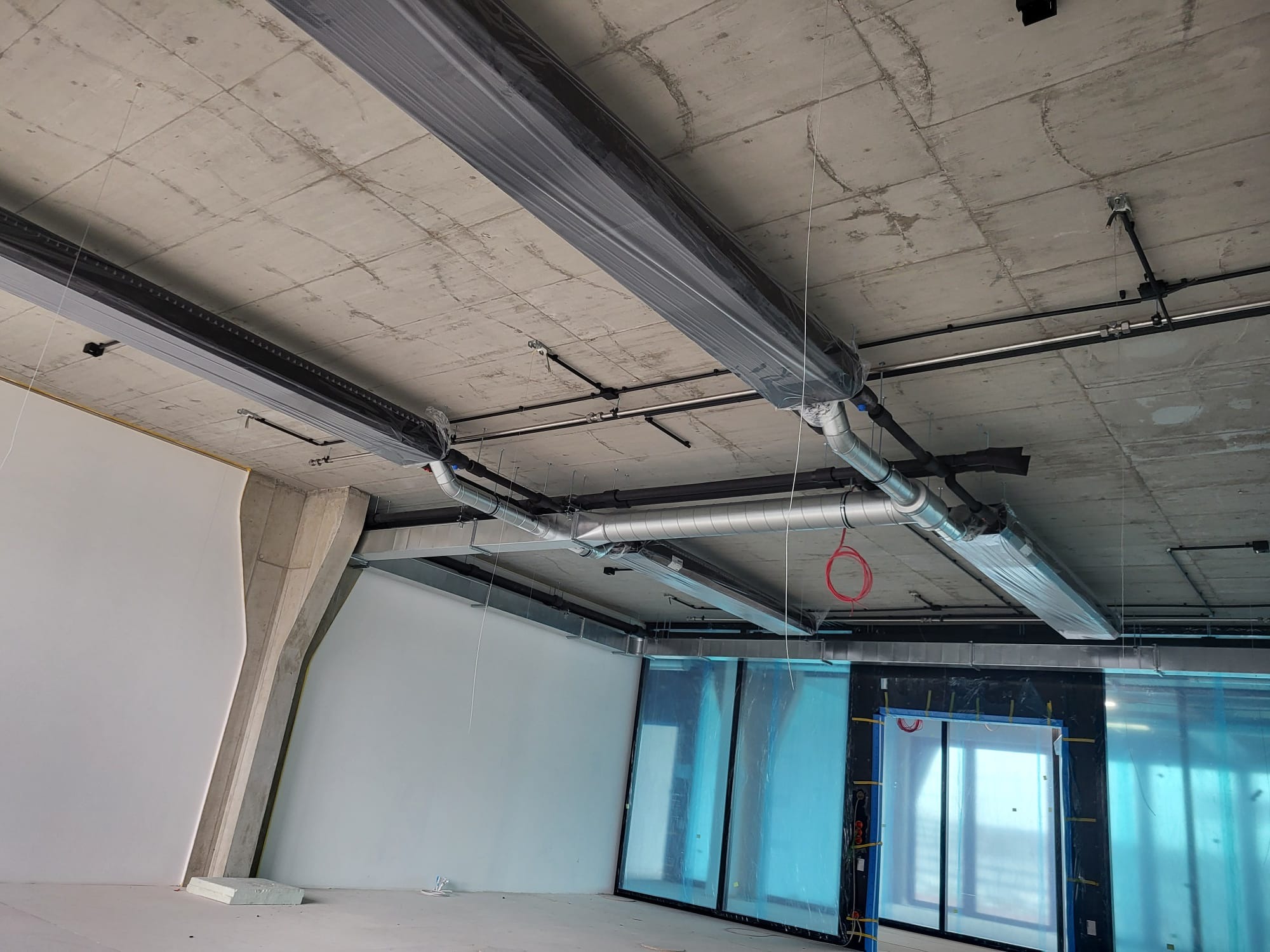Wnętrze w trakcie montażu instalacji wentylacyjnej i hydraulicznej, widoczne rury pod sufitem, zabezpieczone folią elementy oraz surowy betonowy sufit.