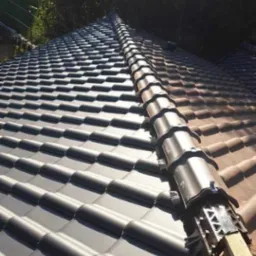 Nowy dach z ciemnej dachówki ceramicznej, widoczne elementy mocujące na kalenicy, w tle zieleń drzew.