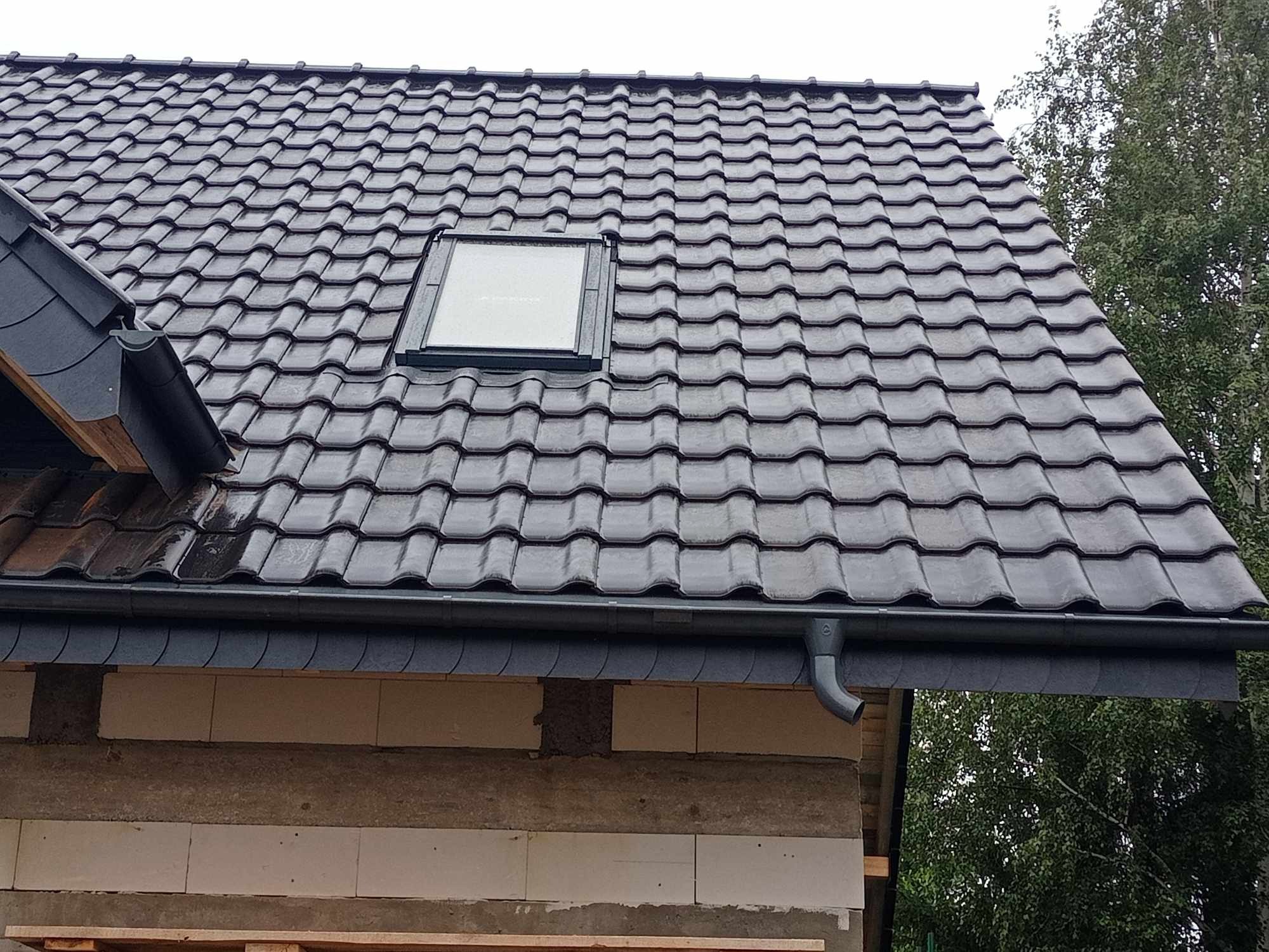 Nowy, ciemny dach z dachówki ceramicznej z oknem dachowym i systemem rynnowym, widok z dołu na fragment budynku w trakcie budowy, z widocznymi bloczkami i drewnianymi elementami konstrukcyjnymi.