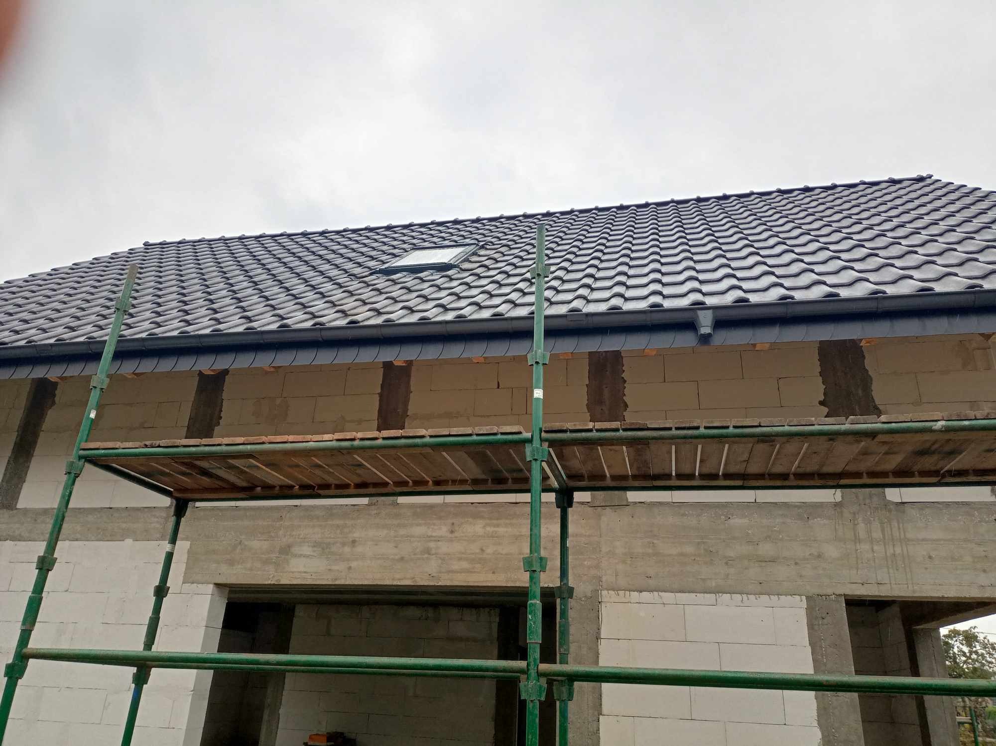 Nowy, grafitowy dach z dachówki ceramicznej z oknem dachowym, widoczny ze stojącego rusztowania na tle surowego stanu budynku z betonu komórkowego.
