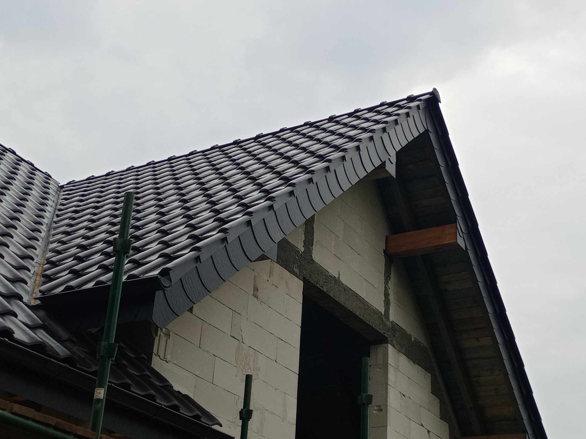Nowy dach z czarnej dachówki karpiówki na budynku w trakcie budowy, widoczne białe bloczki i fragment rusztowania pod pochmurnym niebem.