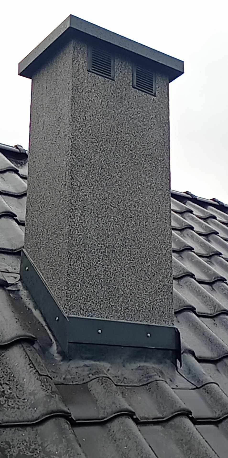 Szary otynkowany komin na dachu pokrytym ciemną dachówką ceramiczną, zabezpieczony obróbką blacharską.