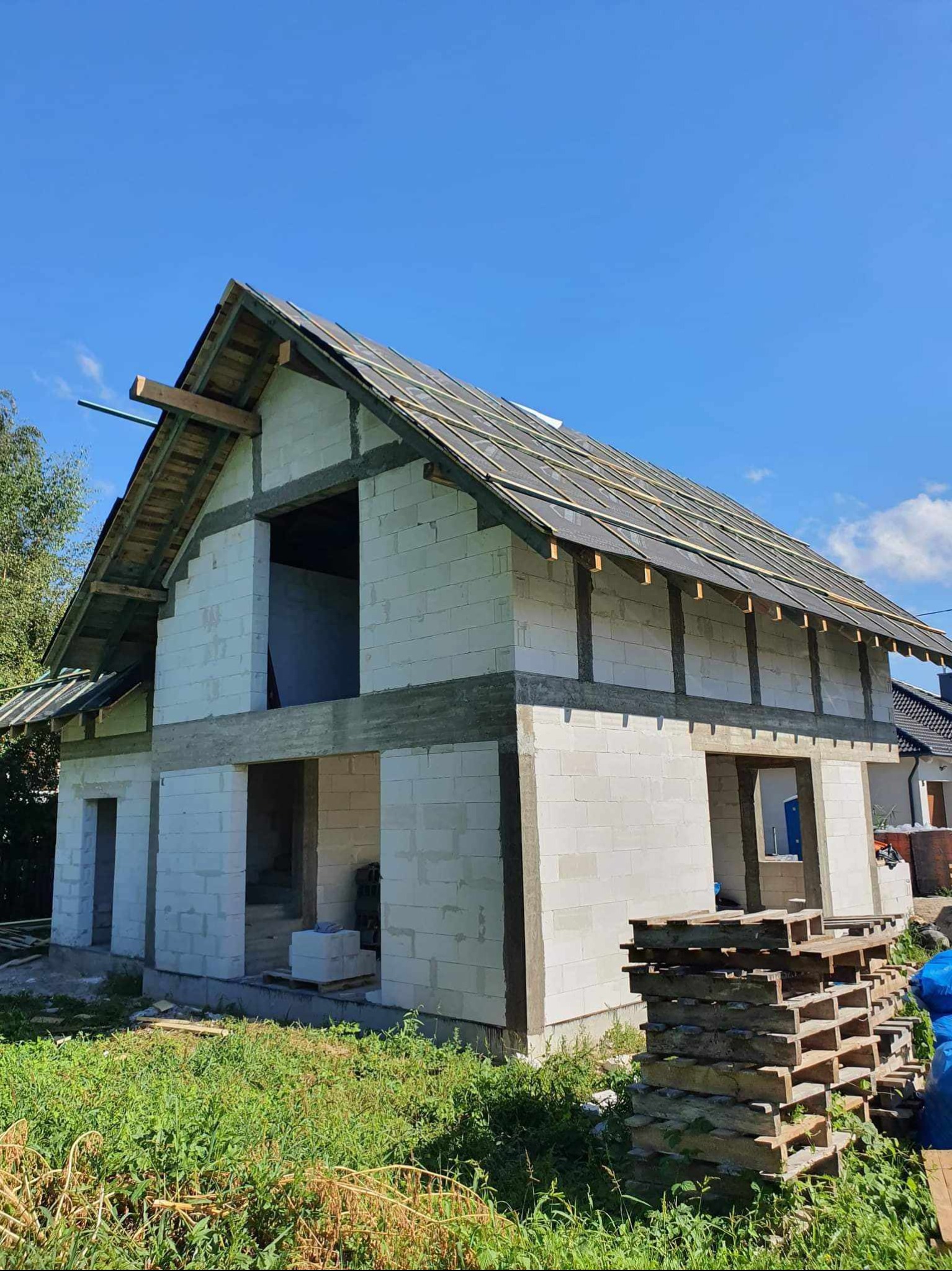 Surowy stan dachu z deskowaniem i papą na domu w trakcie budowy, widoczne białe bloczki konstrukcyjne i palety