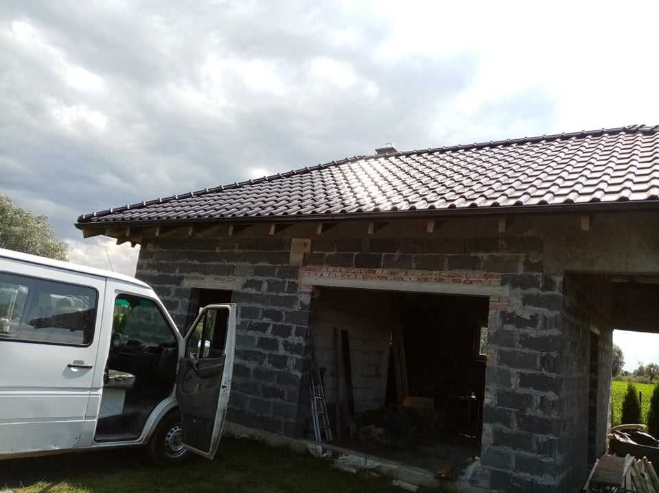 Nowy, brązowy dach z rynnami na surowym budynku z szarych bloczków, obok biały samochód dostawczy z otwartymi drzwiami.