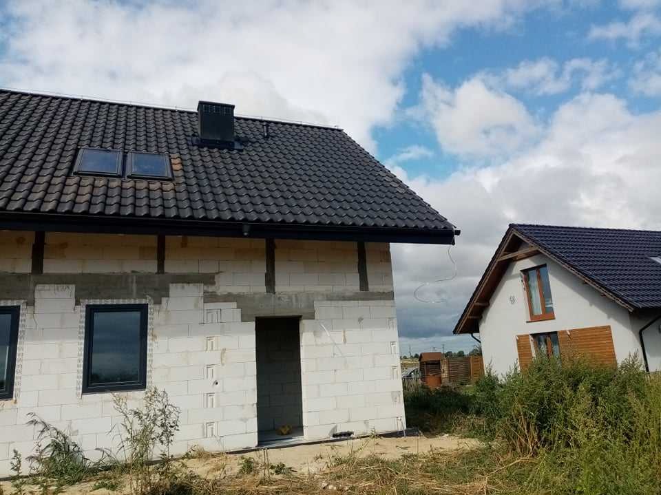 Nowy, ciemny dach z oknami dachowymi na surowym budynku z białego bloczka w Pasłęku, z widocznymi rynnami i kominem, obok drugi budynek z podobnym dachem.