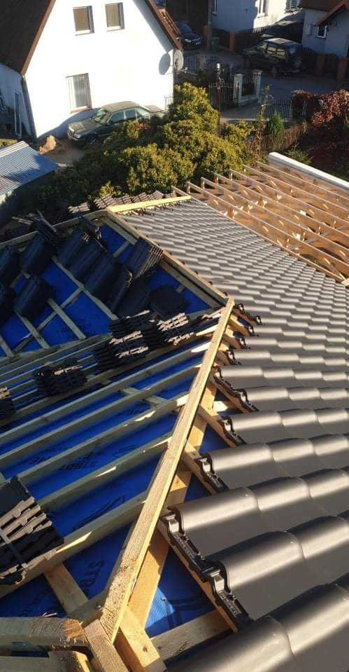 Częściowo pokryty dach z szarą dachówką, widoczna konstrukcja drewniana i niebieska membrana dachowa, w tle dom i drzewa.