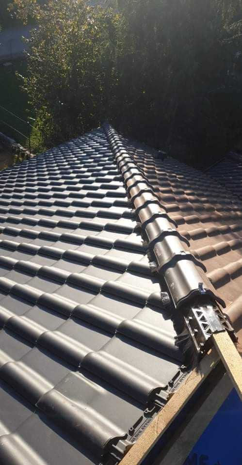 Nowy dach z ciemnej dachówki ceramicznej, widoczne elementy mocujące na kalenicy, w tle zieleń drzew.