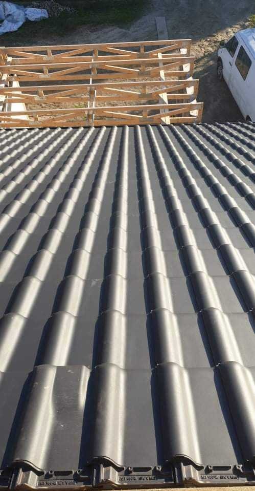 Widok z góry na nowo ułożone, ciemnoszare dachówki ceramiczne z widocznym drewnianym stelażem i fragmentem białego samochodu w tle.