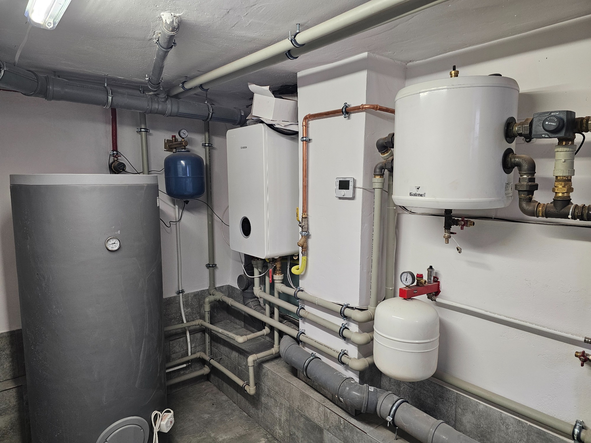 Kompleksowa instalacja grzewcza w kotłowni: zbiornik akumulacyjny, piec gazowy Bosch z otwartym pudełkiem obok, zasobnik CWU Gaimei, liczne rury PCV i miedziane z zaworami.