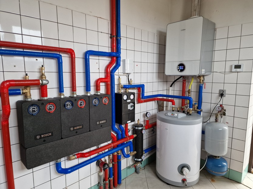 Kompletna, nowoczesna instalacja grzewcza z piecem gazowym, zasobnikiem ciepłej wody i rozdzielaczami podłogówki marki Bosch, rury w izolacji czerwonej i niebieskiej, na tle ściany z białych płytek.