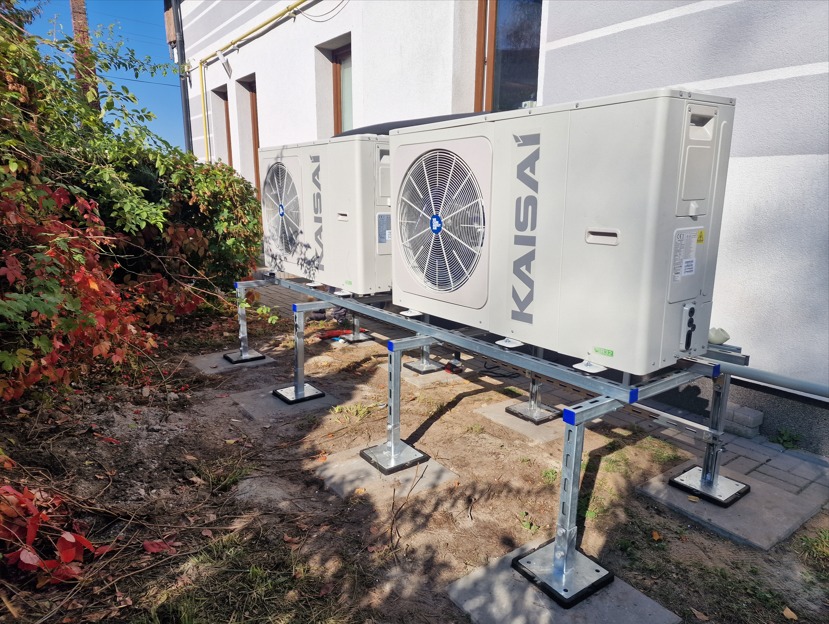 Dwa klimatyzatory marki Kaisai zamontowane na metalowej konstrukcji wsporczej przy ścianie budynku, widoczne elementy instalacji i otoczenie z roślinnością.