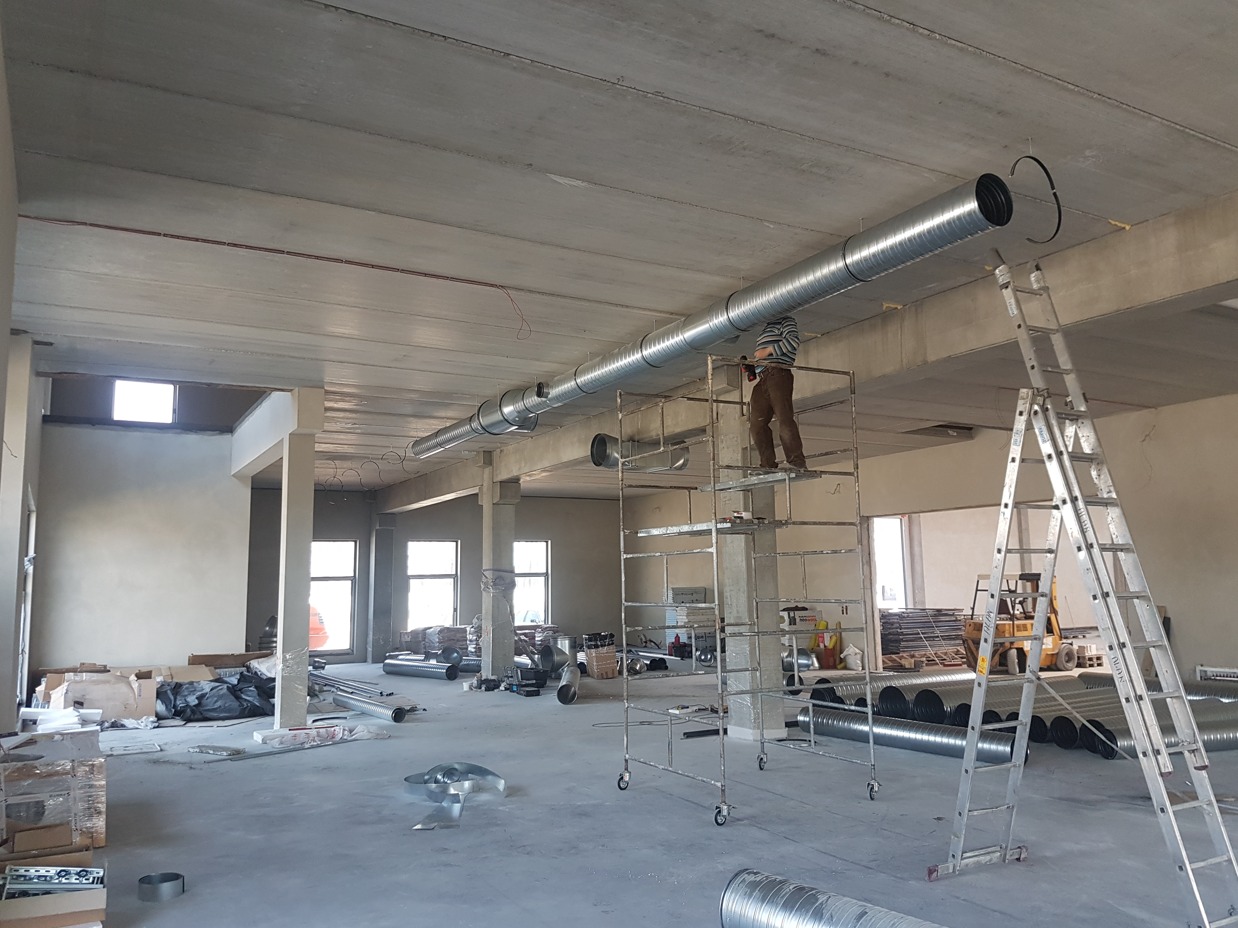 Instalacja wentylacji: mężczyzna na rusztowaniu montuje spiralny przewód wentylacyjny pod betonowym stropem w surowym wnętrzu budynku.