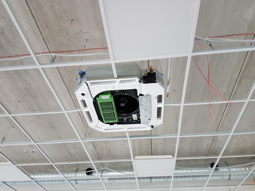 Kasetonowa jednostka klimatyzacji z zielonym filtrem, zamontowana w rastrze sufitowym, widok od dołu, z widocznymi przewodami i elementami instalacji.
