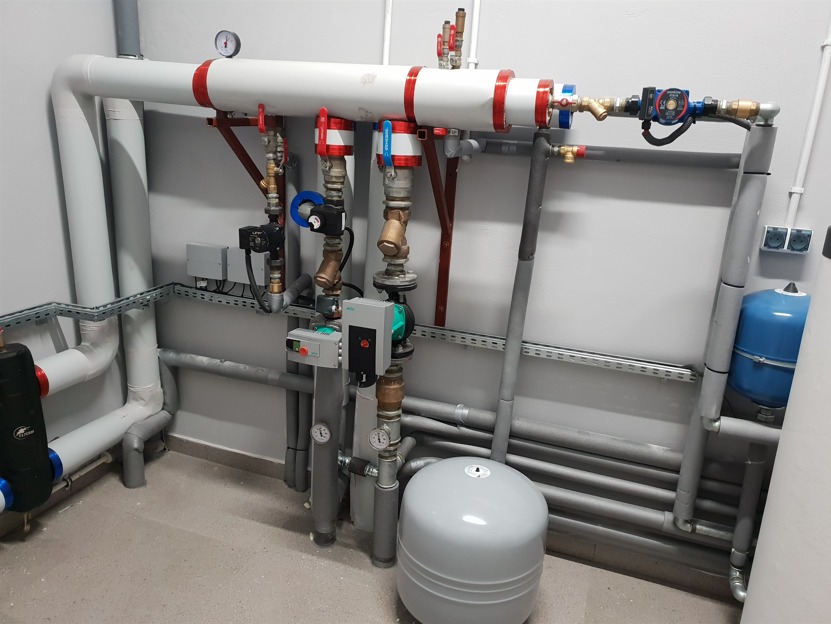 Węzeł cieplny z licznymi rurami, zaworami, pompami i zbiornikami wyrównawczymi w szarej kotłowni, widok z dołu.