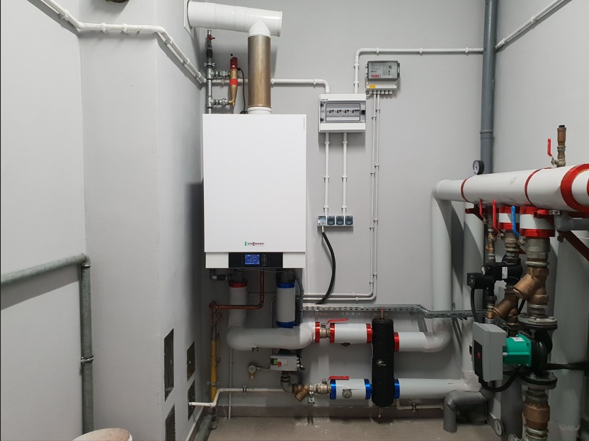 Nowoczesny, biały piec gazowy Viessmann w kotłowni z szarymi ścianami, widoczne rury miedziane i plastikowe, rozdzielnica elektryczna oraz elementy instalacji CO.