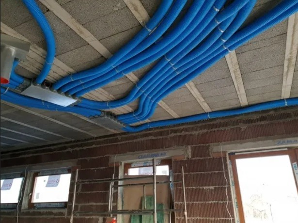 Niebieskie rury instalacji ogrzewania podłogowego zamontowane pod sufitem w surowym stanie, widoczne skrzynki rozdzielcze i fragment ściany z oknami w trakcie budowy.