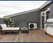 Biała jednostka zewnętrzna klimatyzacji na drewnianym tarasie z szarą ścianą, meblami ogrodowymi i grillem, widok z boku.