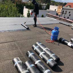 PAWEŁ WIŚNIEWSKI - Pracownik w czarnym ubraniu, z kapturem na głowie, używa palnika do zgrzewania papy na płaskim dachu, obok leżą rolki papy i niebieska butla gazowa.