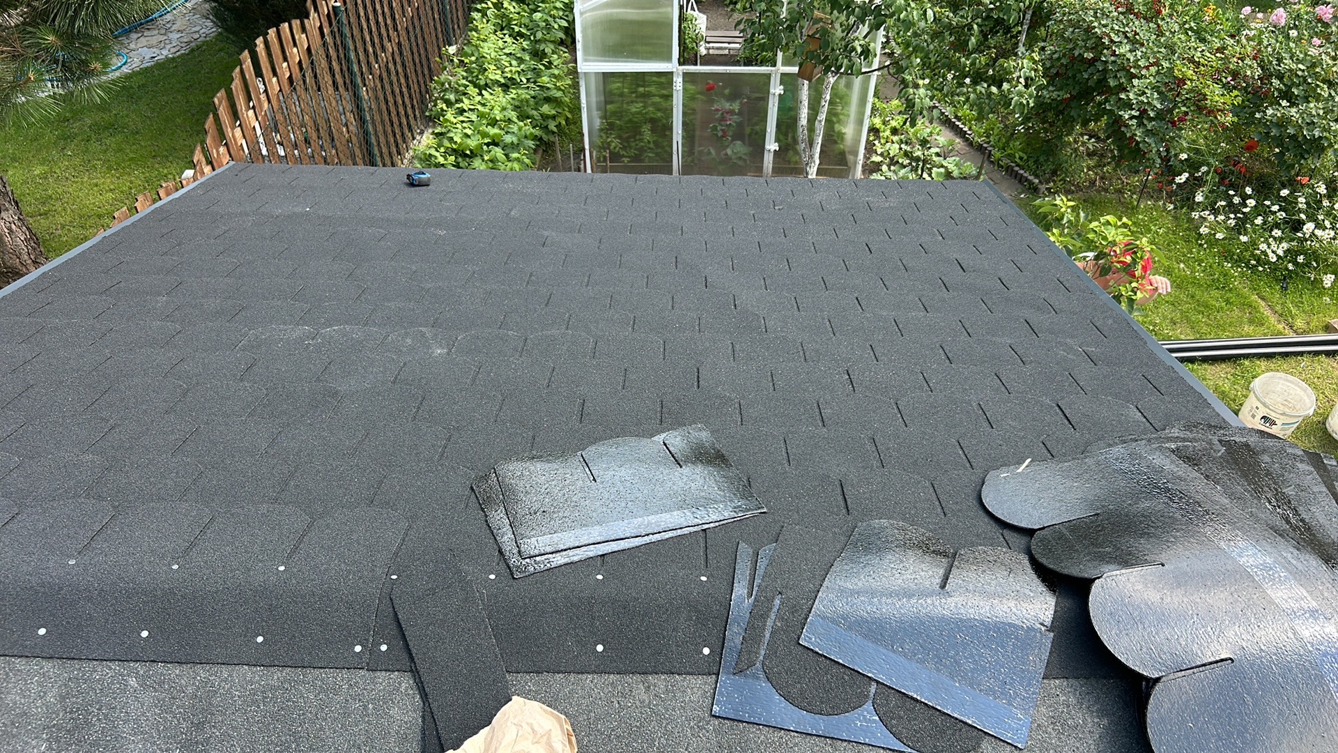 Fragment dachu pokrytego czarnym gontem bitumicznym; widoczne arkusze gontu, wiadro i elementy wykończeniowe na tle zielonego ogrodu z drzewami i szklarnią.