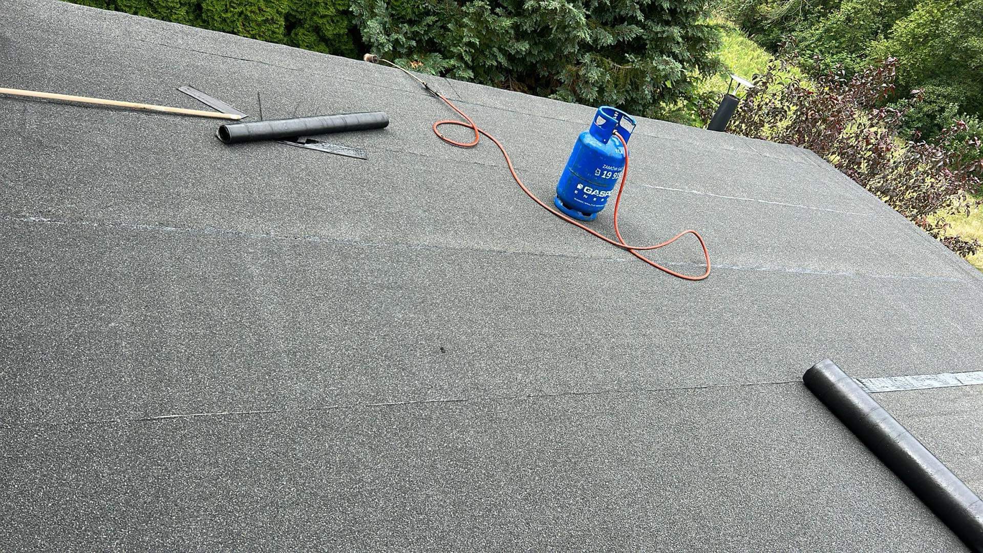 Widok z góry na dach pokryty papą termozgrzewalną, z widoczną butlą gazową, palnikiem dekarskim i narzędziami, w tle zieleń drzew.