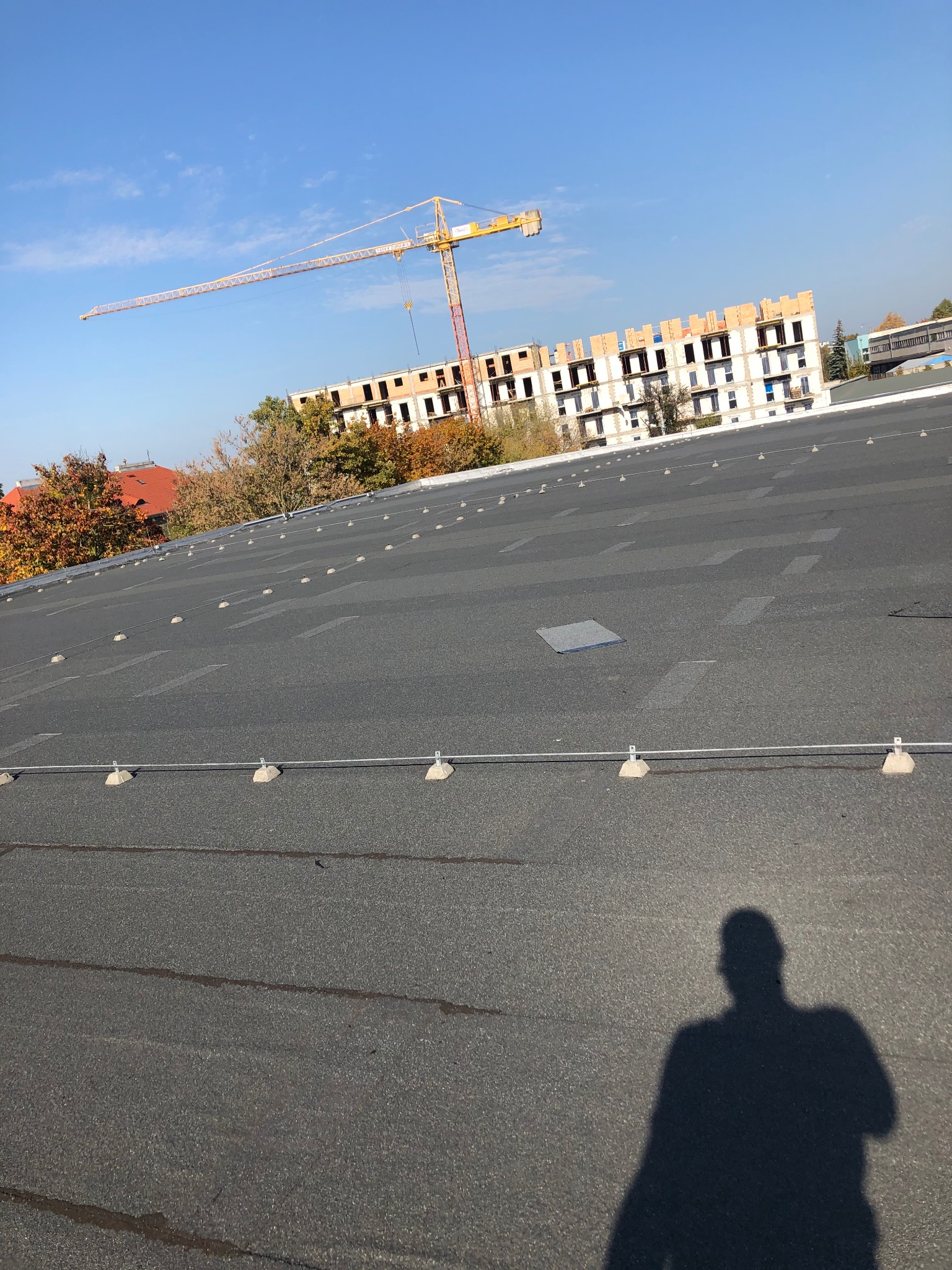 Czarny, płaski dach z widocznymi elementami instalacji odgromowej, w tle budowa nowego budynku z żurawiem i jesienne drzewa, cień osoby w dolnym rogu.