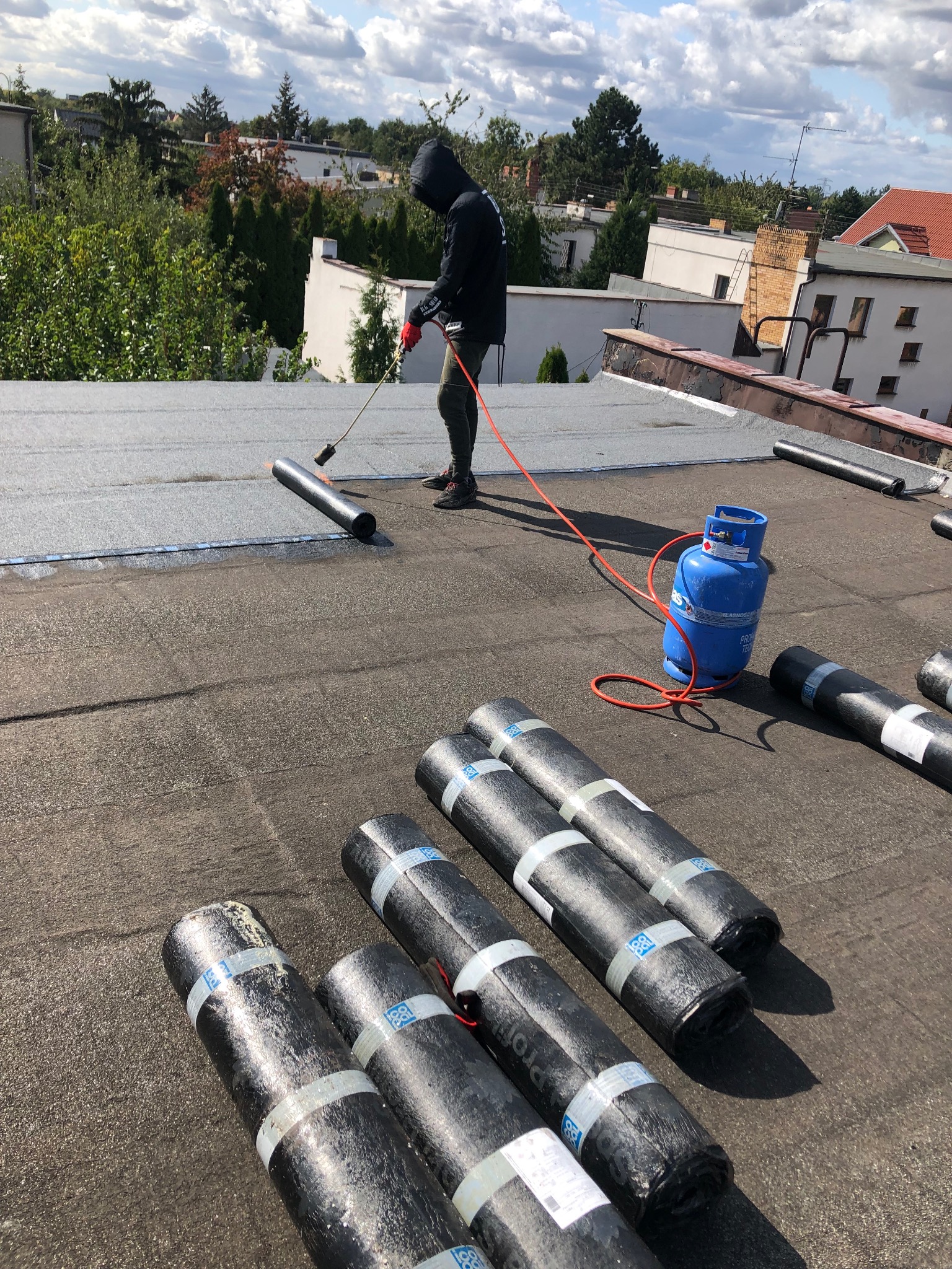 Pracownik w czarnym ubraniu, z kapturem na głowie, używa palnika do zgrzewania papy na płaskim dachu, obok leżą rolki papy i niebieska butla gazowa.